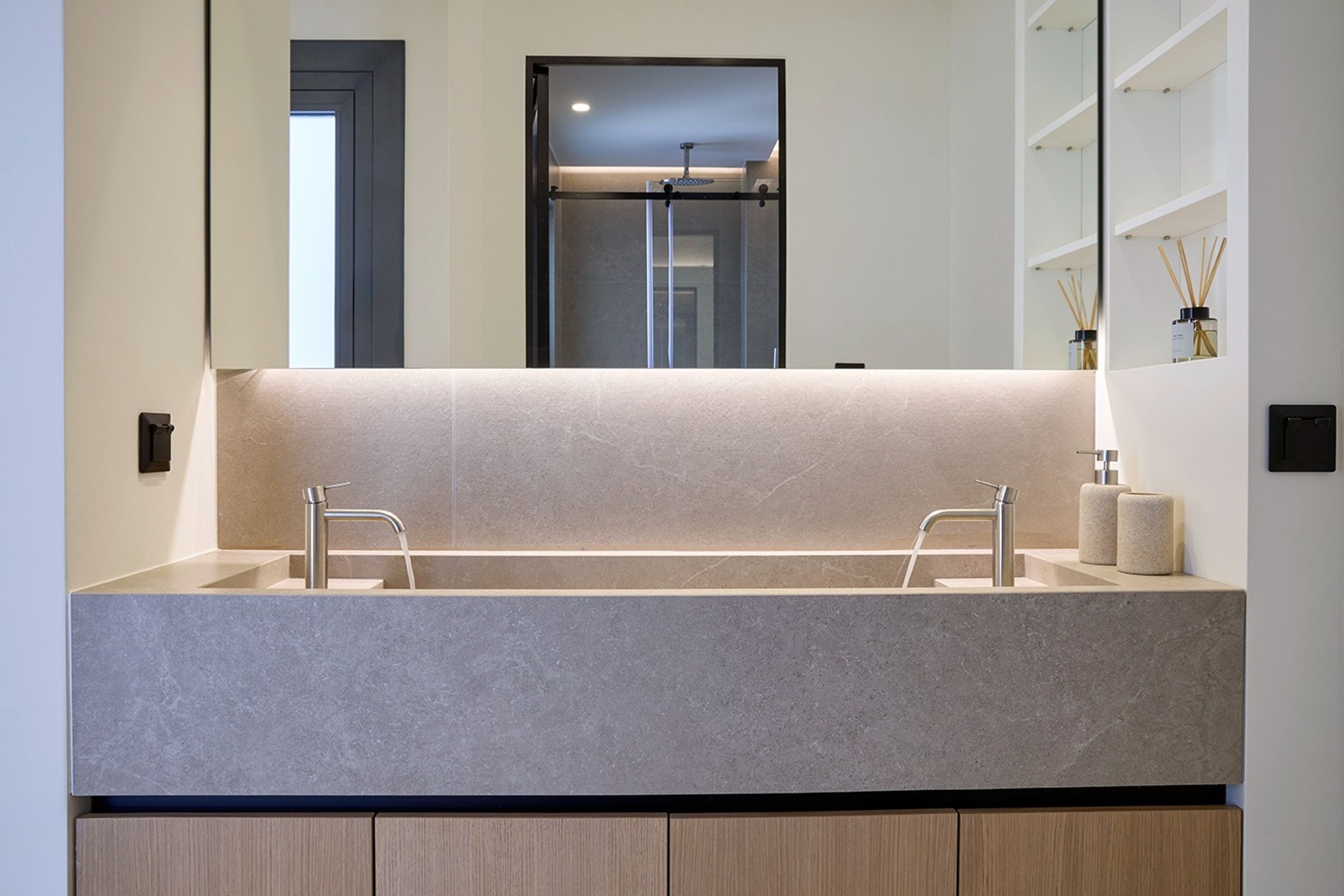 A modern bathroom with a large mirror, two sinks, and a built-in shelf with toiletries.