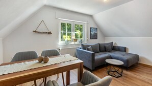 A modern loft bedroom with a sloped ceiling, a large window, a gray sofa, a wooden dining table, and a small round table. - Apartment 'Gellinghausen' with Shared Terrace, Private Garden and Wi-Fi (Schmallenberg)