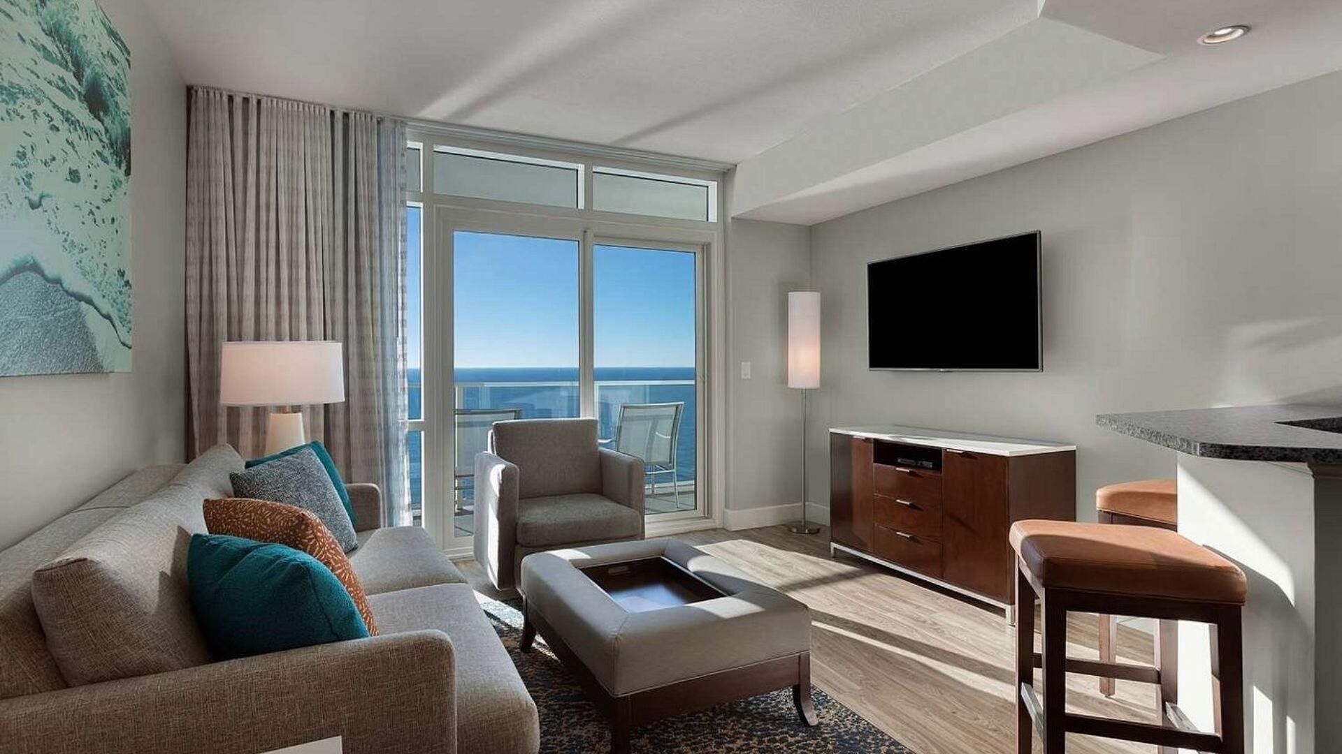 A modern hotel room with a large window offering a view of the ocean, a flat-screen TV mounted on the wall, a sofa with multiple cushions, a small coffee table, and a bar area with stools.
