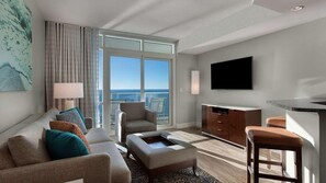 A modern hotel room with a large window offering a view of the ocean, a flat-screen TV mounted on the wall, a sofa with multiple cushions, a small coffee table, and a bar area with stools.