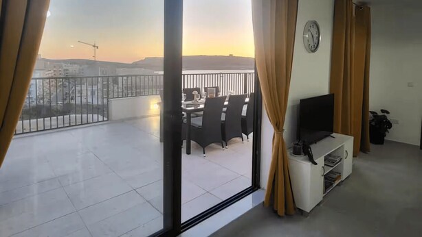 A balcony with a dining table and chairs, a TV on a stand, and curtains. - Unique Waterfront Penthouse: Lovely Sea Views - Spacious Balcony! (Il-Mellieħa)