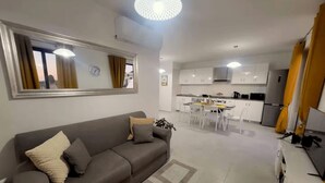 A modern living room with a grey sofa, a dining area with white chairs, and a kitchen with white cabinets. - Unique Waterfront Penthouse: Lovely Sea Views - Spacious Balcony! (Il-Mellieħa)