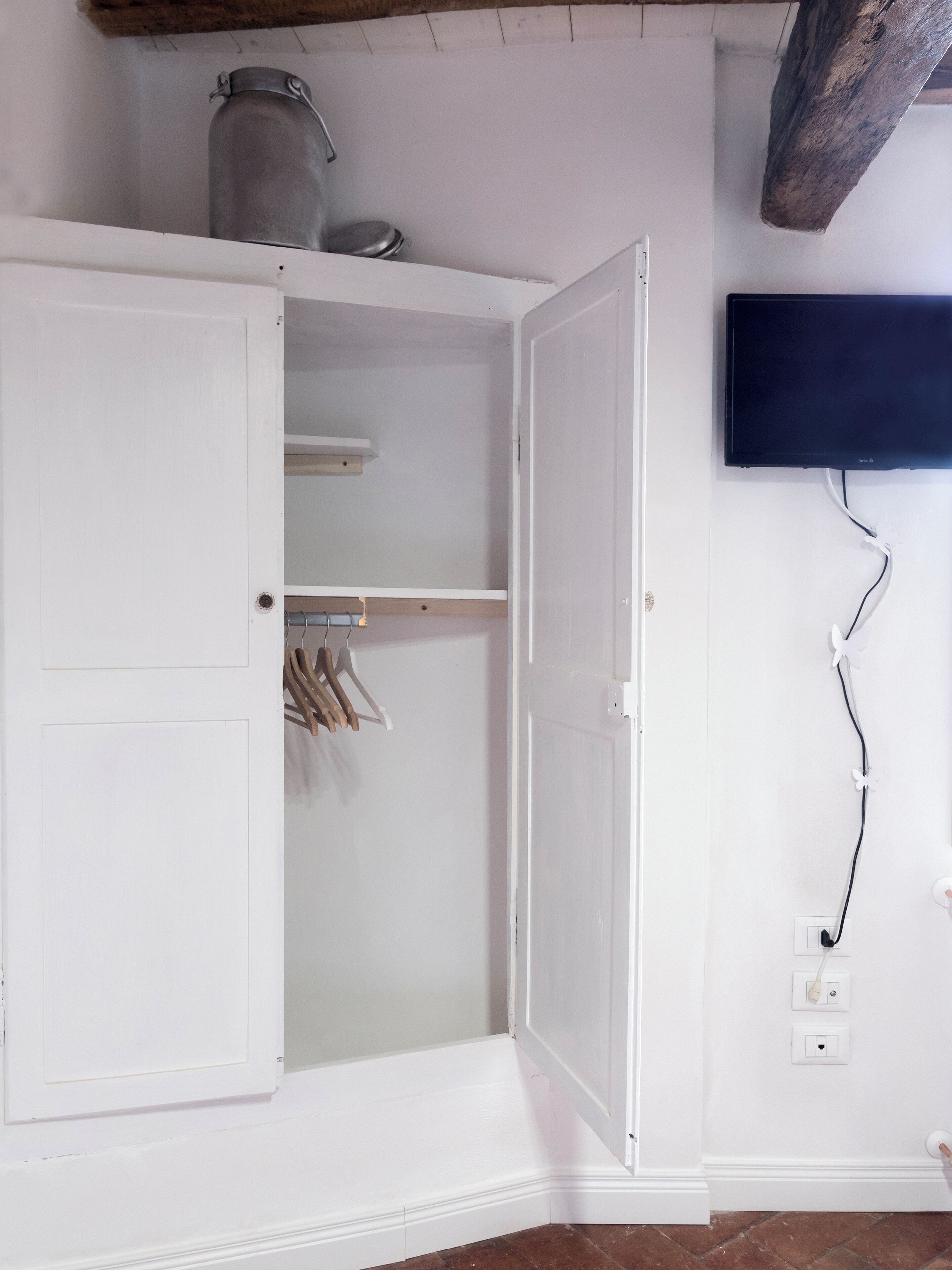A room with a white wardrobe, a hanging rack, a television mounted on the wall, and a power outlet.