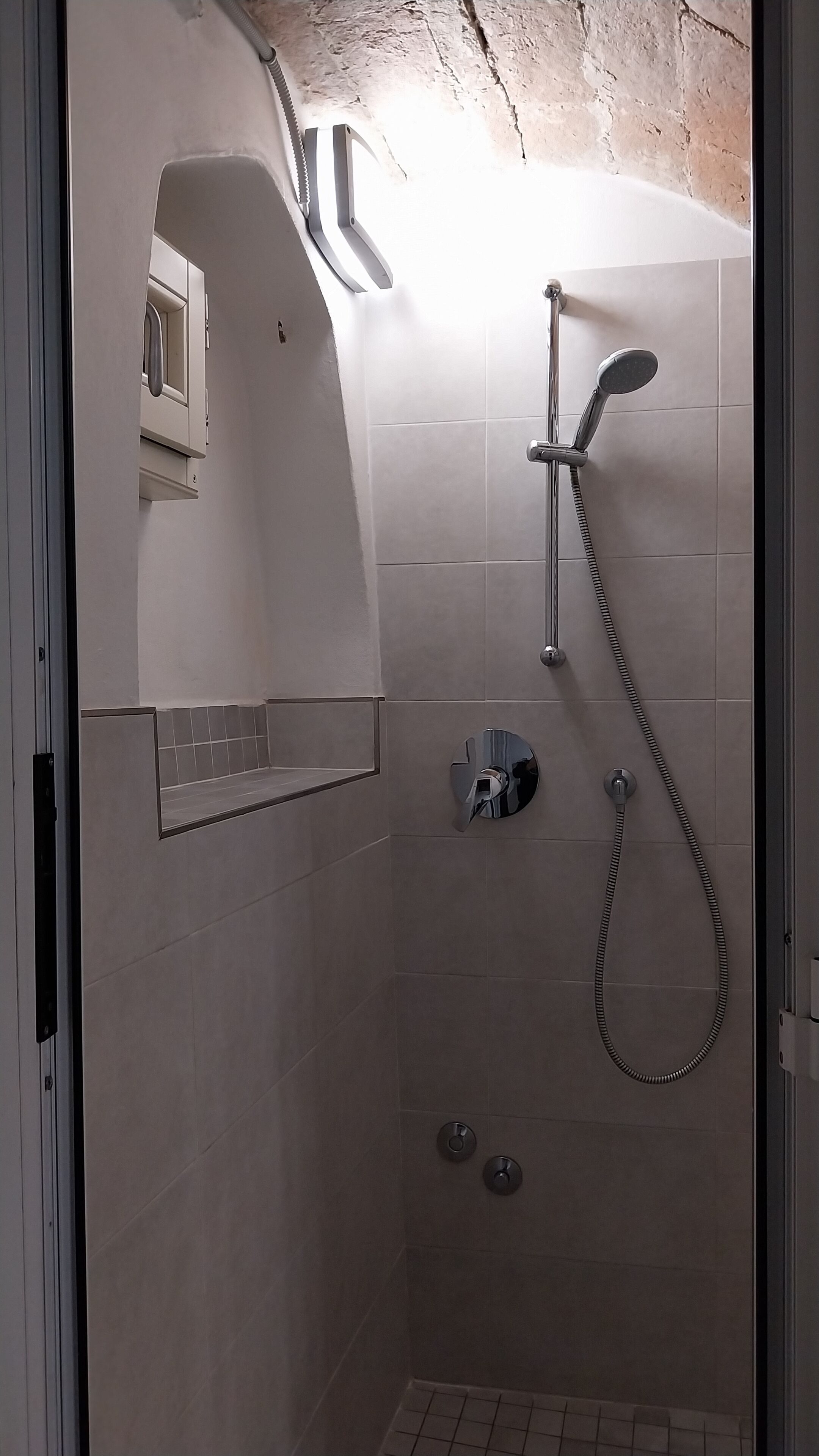 A shower with a handheld showerhead and a wall-mounted faucet, a small shelf, and a window with a towel hanging out.