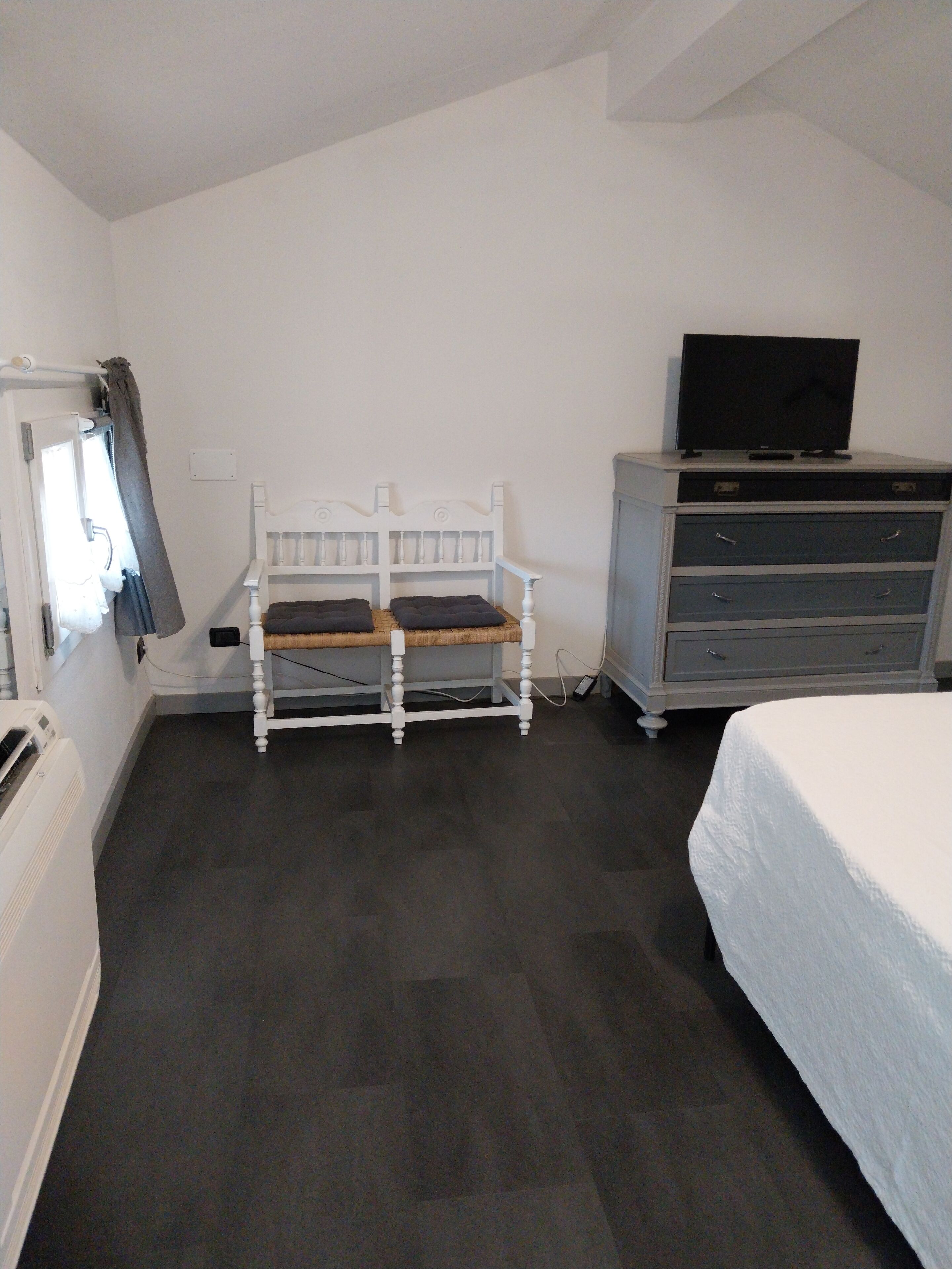 A room with a bed, a white dresser, a television on top, and a chair with cushions.