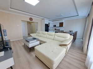 A modern living room with a sectional sofa, a coffee table, and a TV stand. There is a kitchen area in the background with cabinets and a dining table.