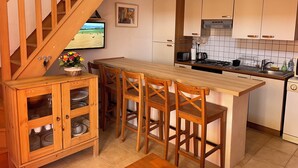 A kitchen with wooden cabinets, a wooden dining table with chairs, a TV on the wall, and a painting.