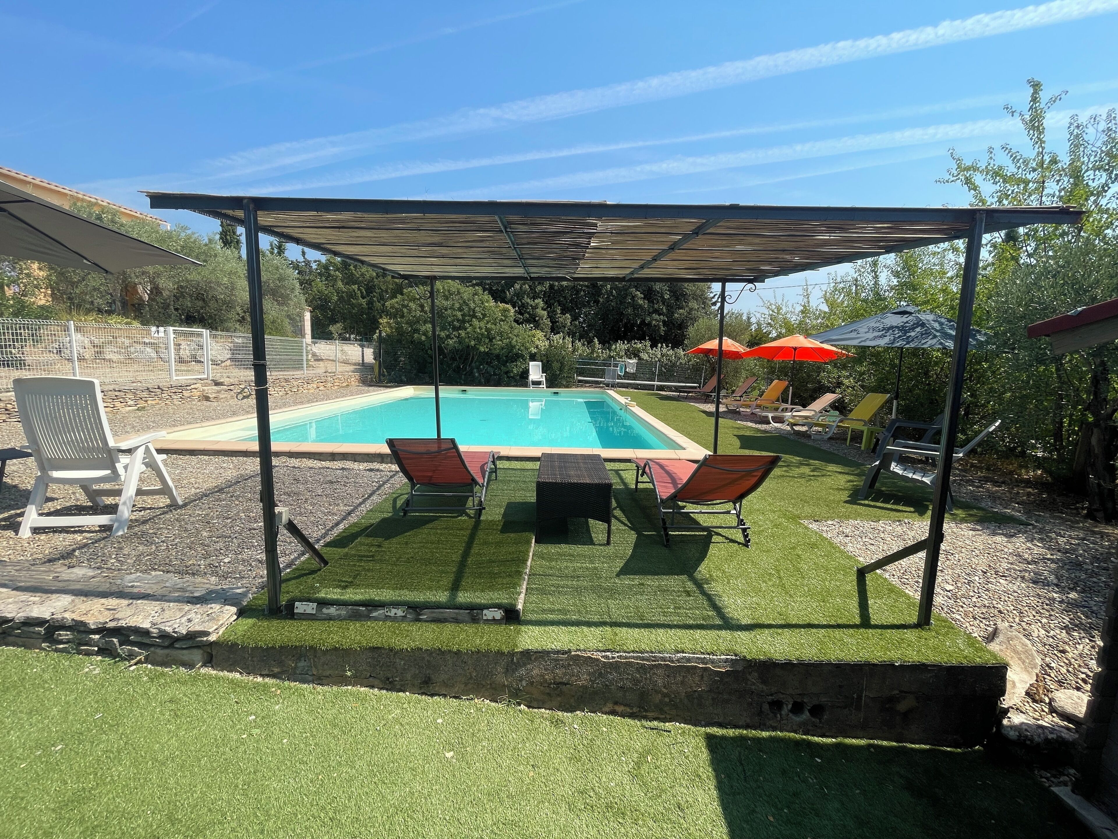 Patio con piscina, césped artificial y mobiliario exterior bajo una pérgola.