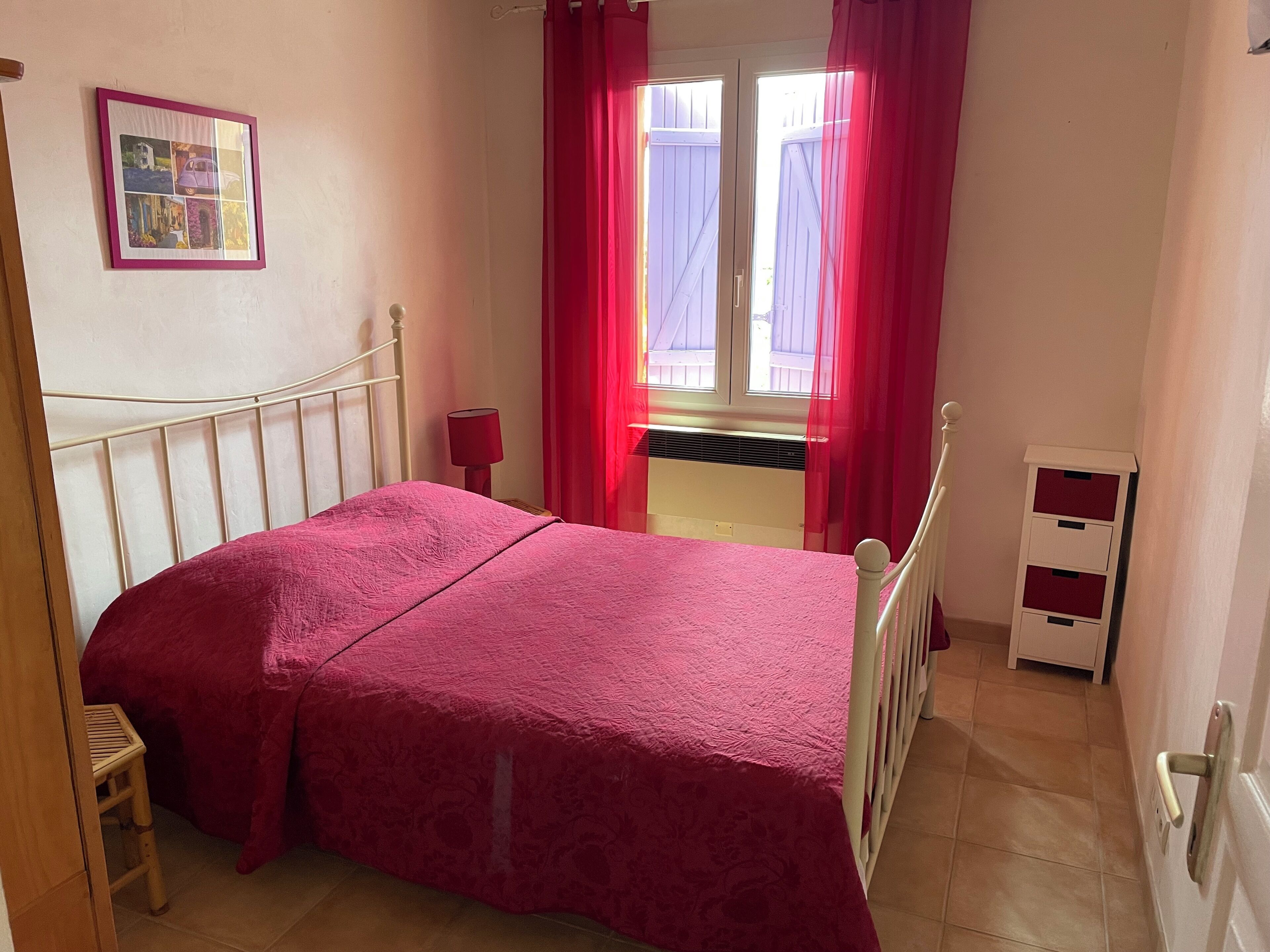 A bedroom with a bed, a bedside table with a lamp, a window with curtains, and a small stool.