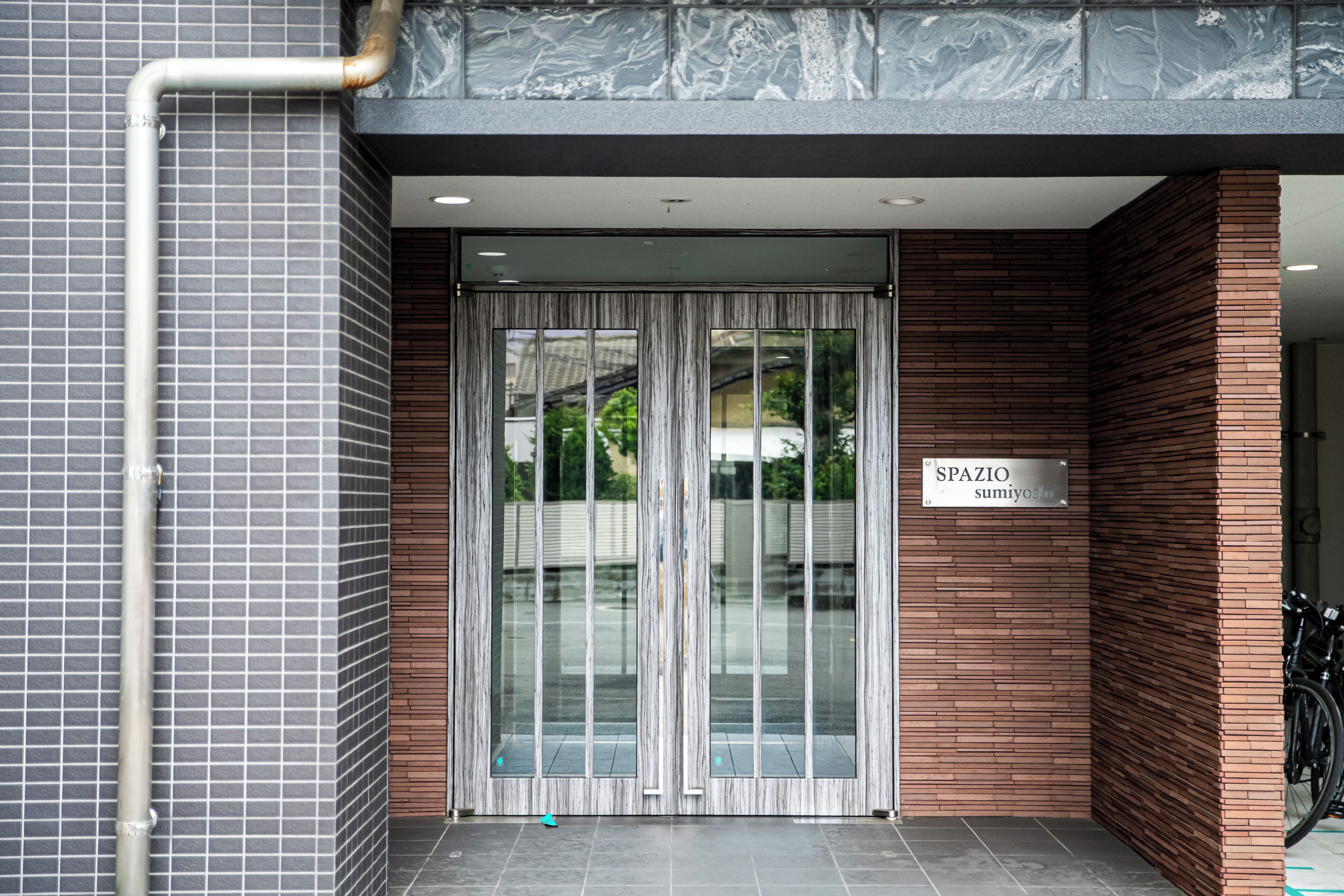 Entrada de un edificio con pared de azulejos, una tubería de metal, una puerta de vidrio con cortina y un cartel que dice 'SPAZIO sumiyoshi'.