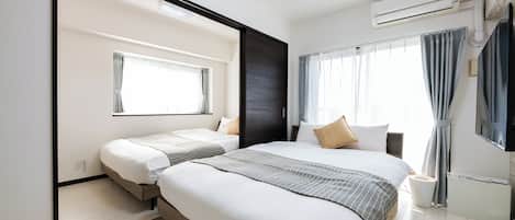 Una habitación de hotel moderna con una cama grande, una cama más pequeña, un televisor de pantalla plana, un equipo de aire acondicionado y una ventana con cortinas.