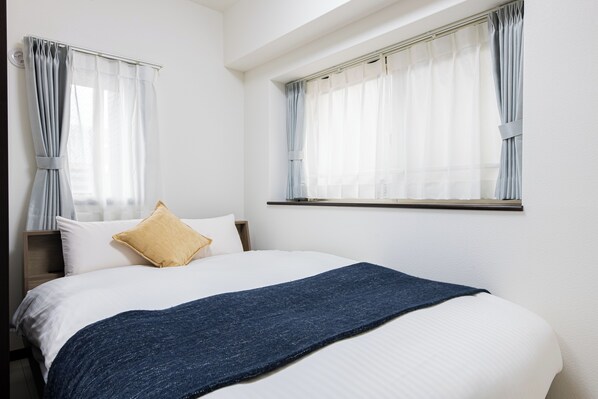 Un dormitorio con una cama, dos ventanas con cortinas, una mesita de noche y una mesilla auxiliar.