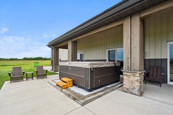 A hot tub outside a building with a covered patio area, two chairs, and a wooden stool.