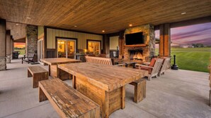 An outdoor patio area with a large wooden table and benches, a stone fireplace, and a television mounted on the wall.