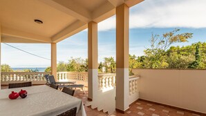 A covered outdoor patio with a table and chairs, a view of the sea, and a balcony railing.