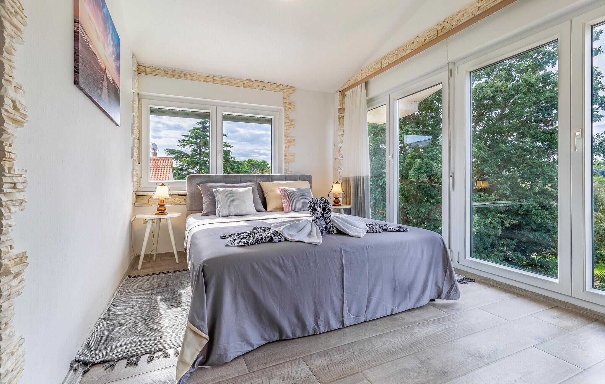 A bedroom with a large bed, two bedside tables with lamps, a view of greenery through large windows, and a stone wall on one side.