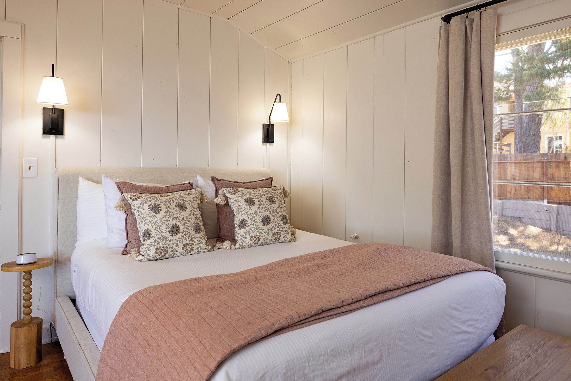 A bedroom with a large bed, two wall-mounted lamps, a bedside table, and a window with curtains.