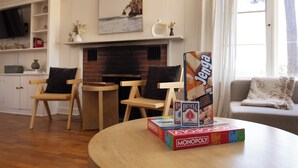 A living room with a fireplace, a bookshelf, and a table with Monopoly and Bicycle cards. - Charming Carmel Cottage Ocean & Mntn View Walk DT (Carmel)