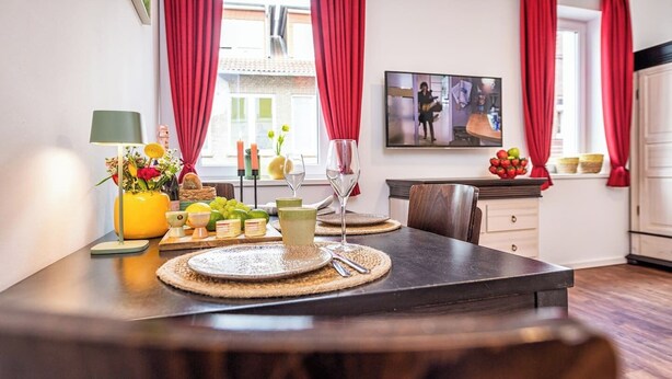 Un área de comedor con una mesa lista para dos personas, un jarrón de flores, velas y un televisor al fondo.