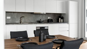Una cocina moderna con armarios blancos, encimera gris y comedor con mesa de madera y sillas negras.