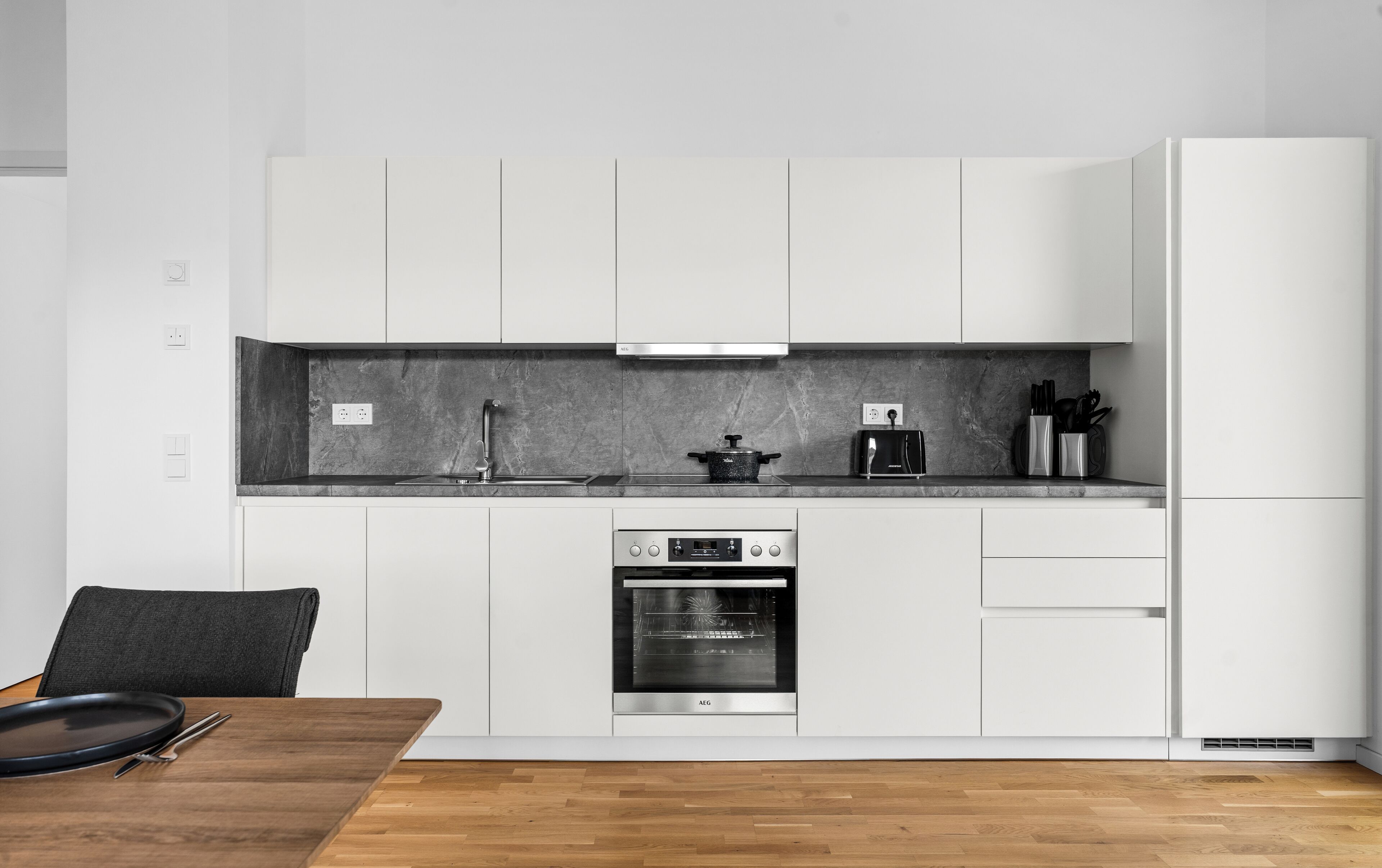 Een moderne keuken met witte kastjes, een ingebouwde oven, een kookplaat en een aanrecht met een pan en keukengerei.
