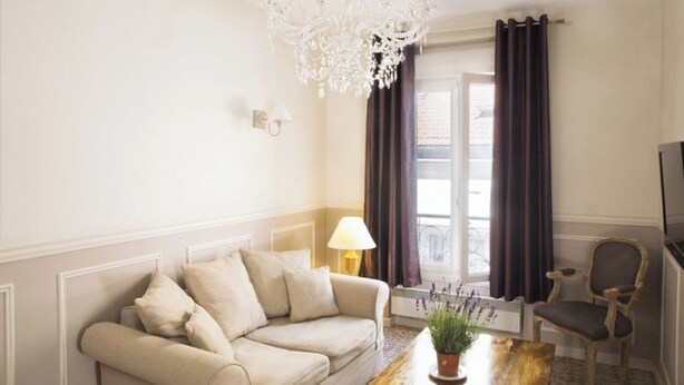 A living room with a beige sofa, a wooden coffee table with a plant, a chandelier, a TV, and a window with curtains. - Practical Apartment for 3 people in charming Paris (Paris)