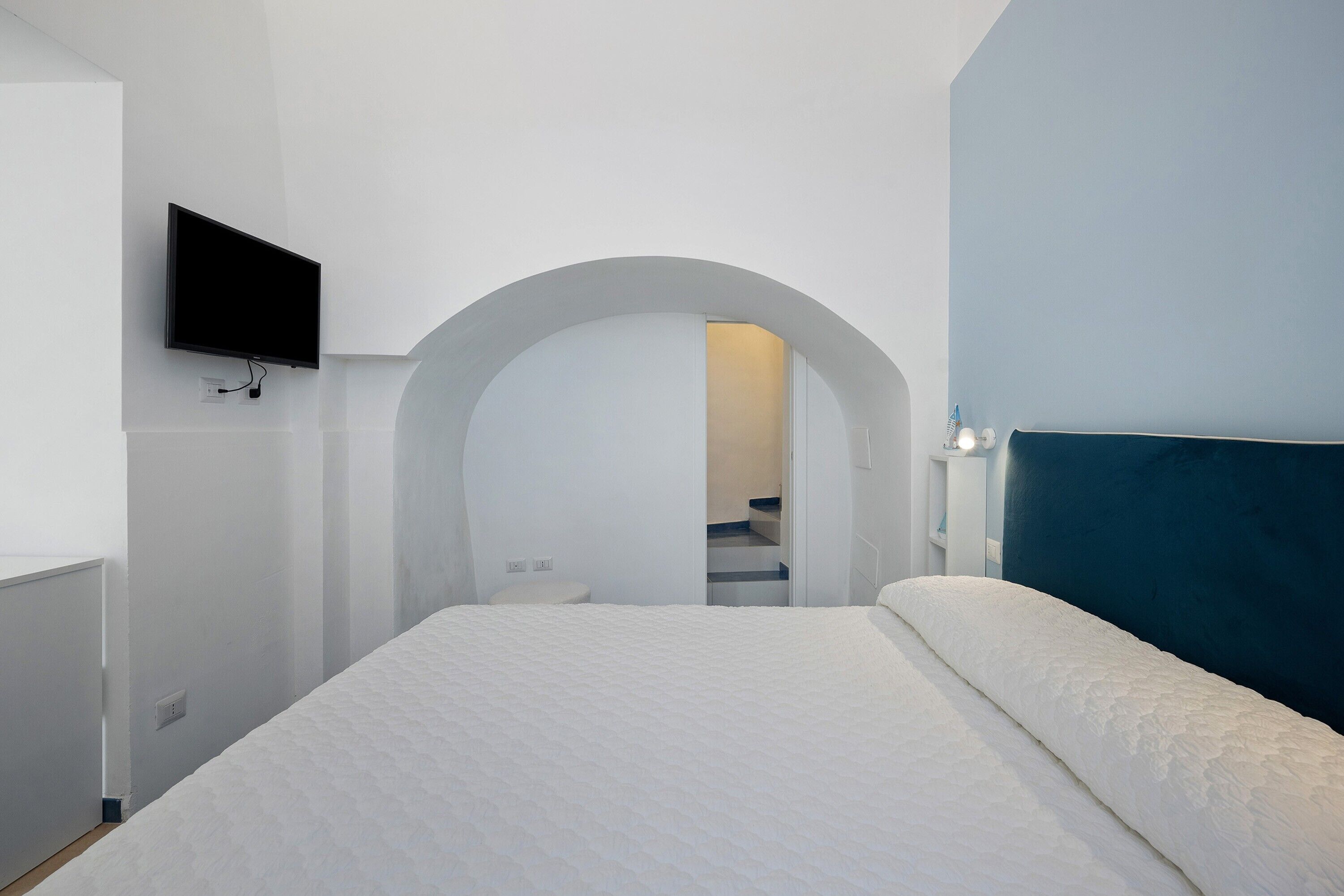 A bedroom with a bed, a TV mounted on the wall, and an arched doorway leading to a bathroom.