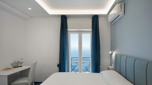 A bedroom with a bed, a desk, and a chair. There is a window with curtains and a view of the outside. - Room 'Camera Furore Sea View' with Sea View, Wi-Fi and Air Conditioning (Furore)