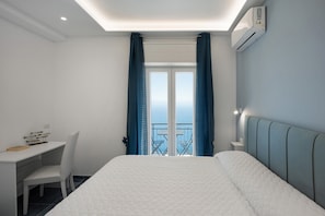A bedroom with a bed, a desk, and a chair. There is a window with curtains and a view of the outside. - Room 'Camera Furore Sea View' with Sea View, Wi-Fi and Air Conditioning (Furore)