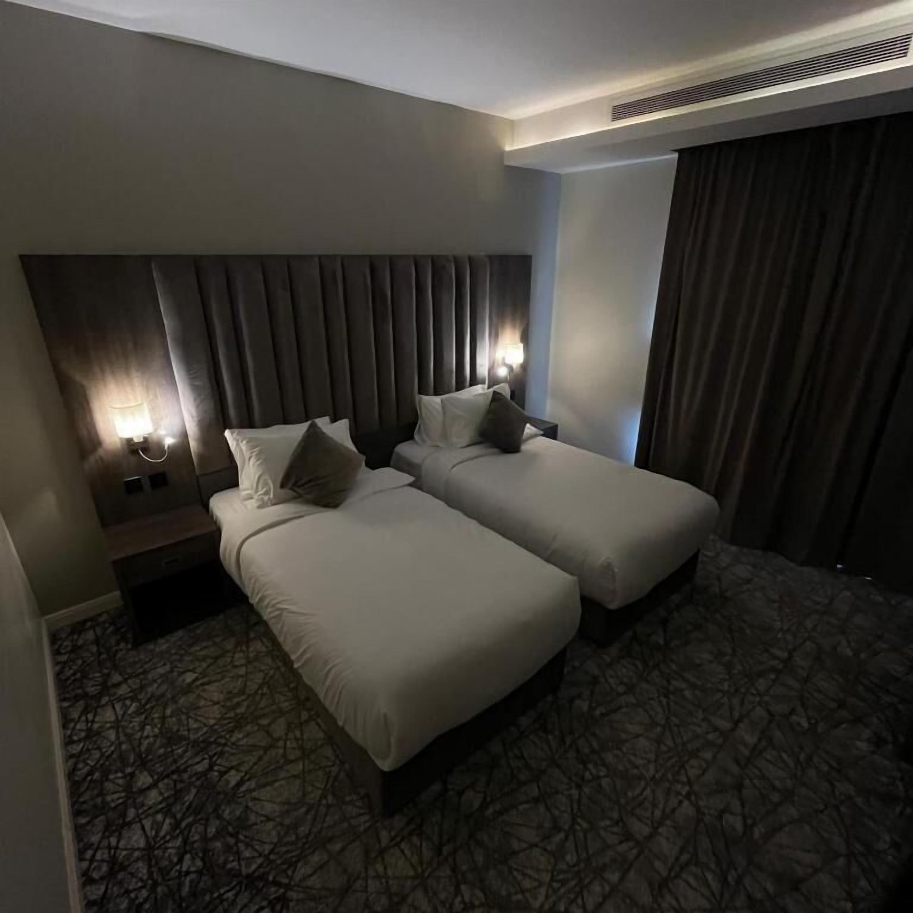Habitación de hotel con dos camas individuales, cabecera con listones verticales, mesita de noche con lámpara y alfombra estampada.