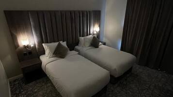 Habitación de hotel con dos camas individuales, cabecera con listones verticales, mesita de noche con lámpara y alfombra estampada.