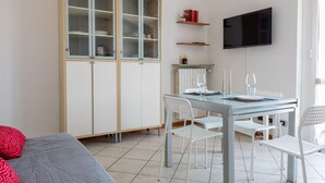 Ett modernt rum med ett matbord med glasskiva, vita stolar, ett skåp med glasdörrar, en platt-TV och en säng med röda kuddar.