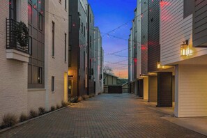 Uma viela estreita com edifícios de tijolo, luzes de corda e céu ao pôr do sol.
