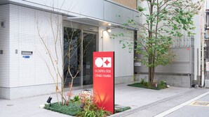 A building with a tiled exterior, a red sign with white text, and a small garden area with plants and a tree. - UCHIWA STAY Osaka NAMBA (Osaka)