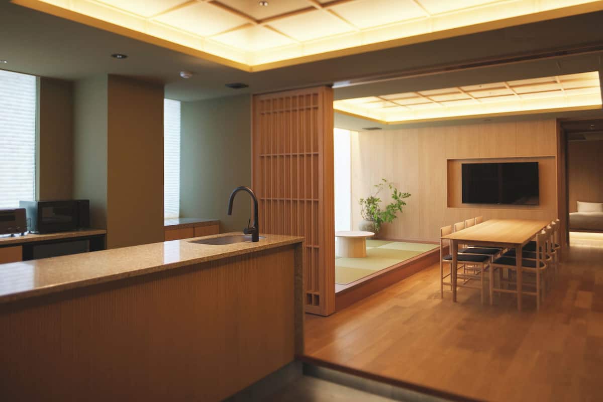 A modern kitchen with a wooden countertop, a sink, and a microwave. There is a dining area with a table and chairs, and a TV mounted on the wall.