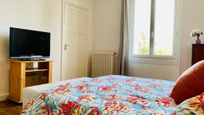 A bedroom with a bed, a television on a wooden stand, a window with curtains, and a radiator. - VILLA COSY (fumel)