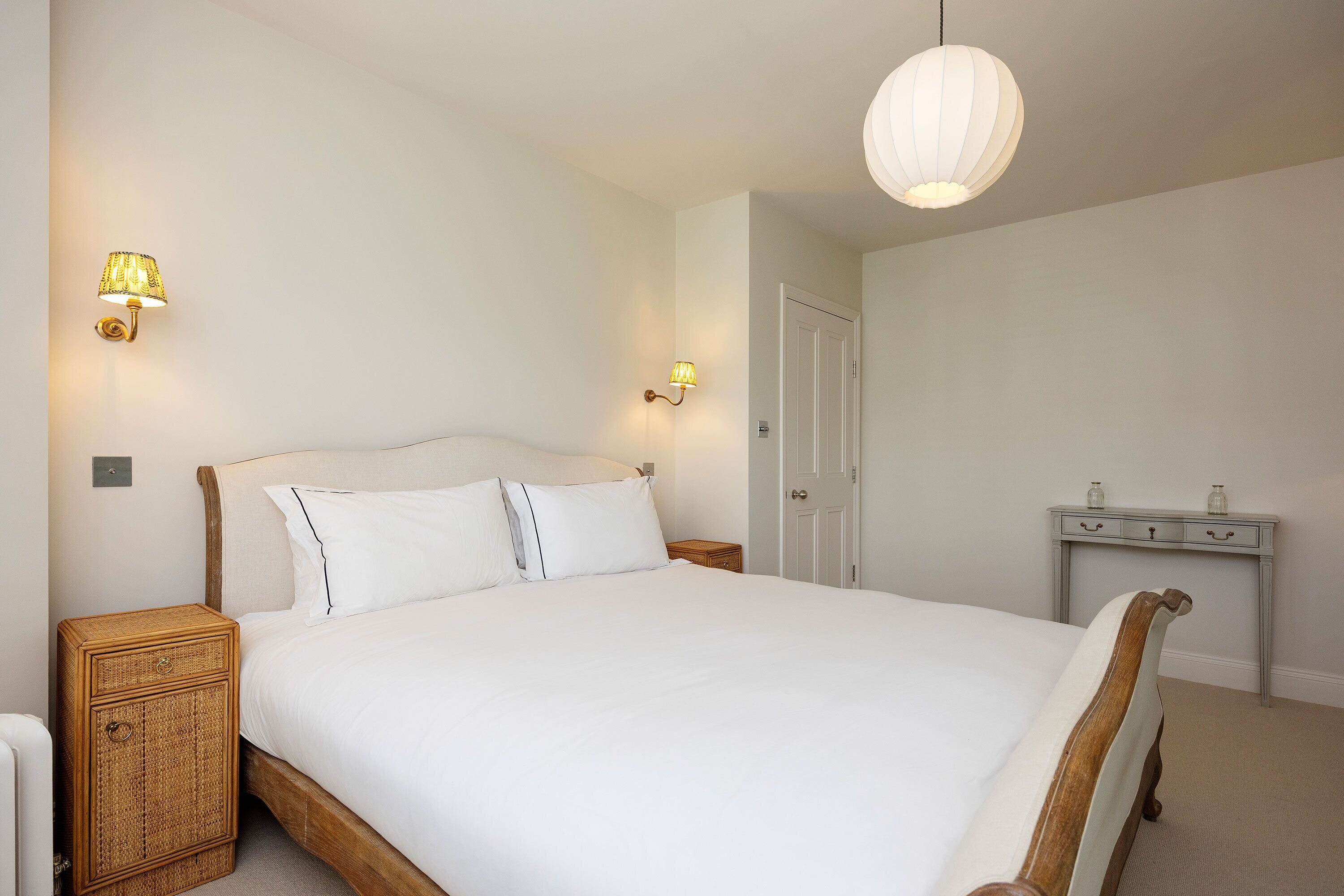 Een keurig opgemaakt bed met witte lakens, twee wandlampen en een hanglamp.