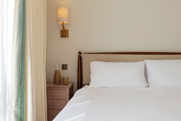 A neatly made bed with white linens, a bedside lamp, a small wicker basket, and a wooden nightstand with drawers.