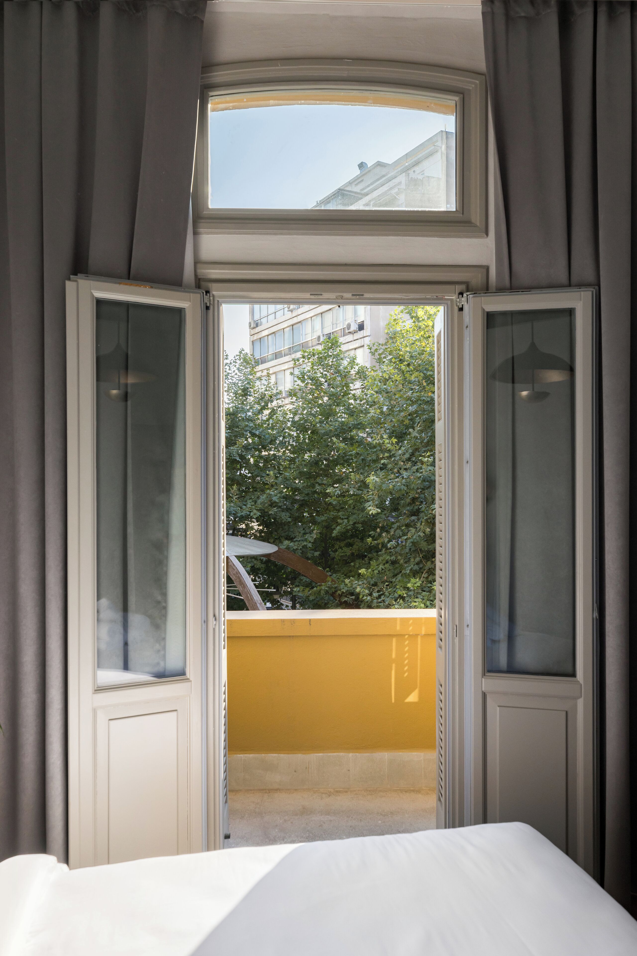 Ein Schlafzimmer mit großem Fenster, Vorhängen und Blick auf ein gelbes Gebäude und Bäume.