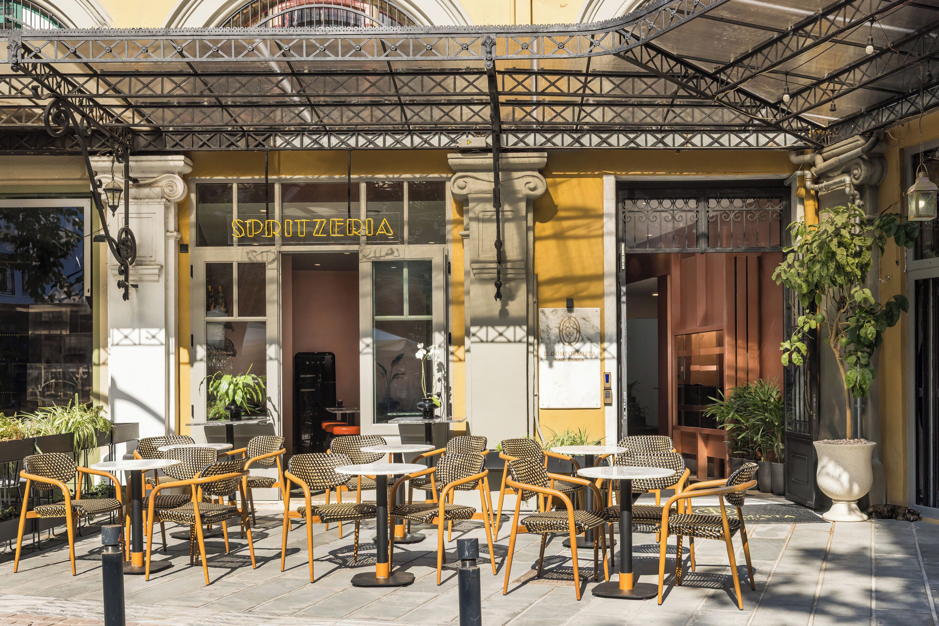 Ein Straßencafé mit Korbstühlen und Tischen, einer gelben Fassade und einem Schild mit der Aufschrift 'SPRITZFRIA'.