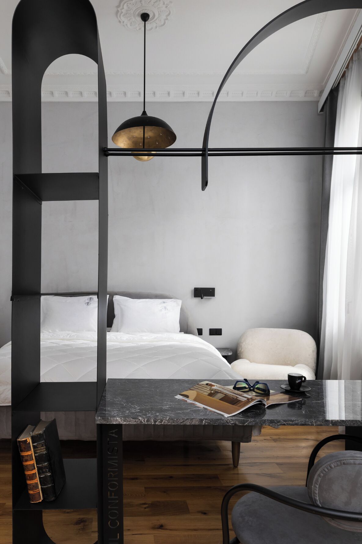 A modern hotel room with a bed, a desk with a magazine and glasses, a chair, and a unique black and white decor.