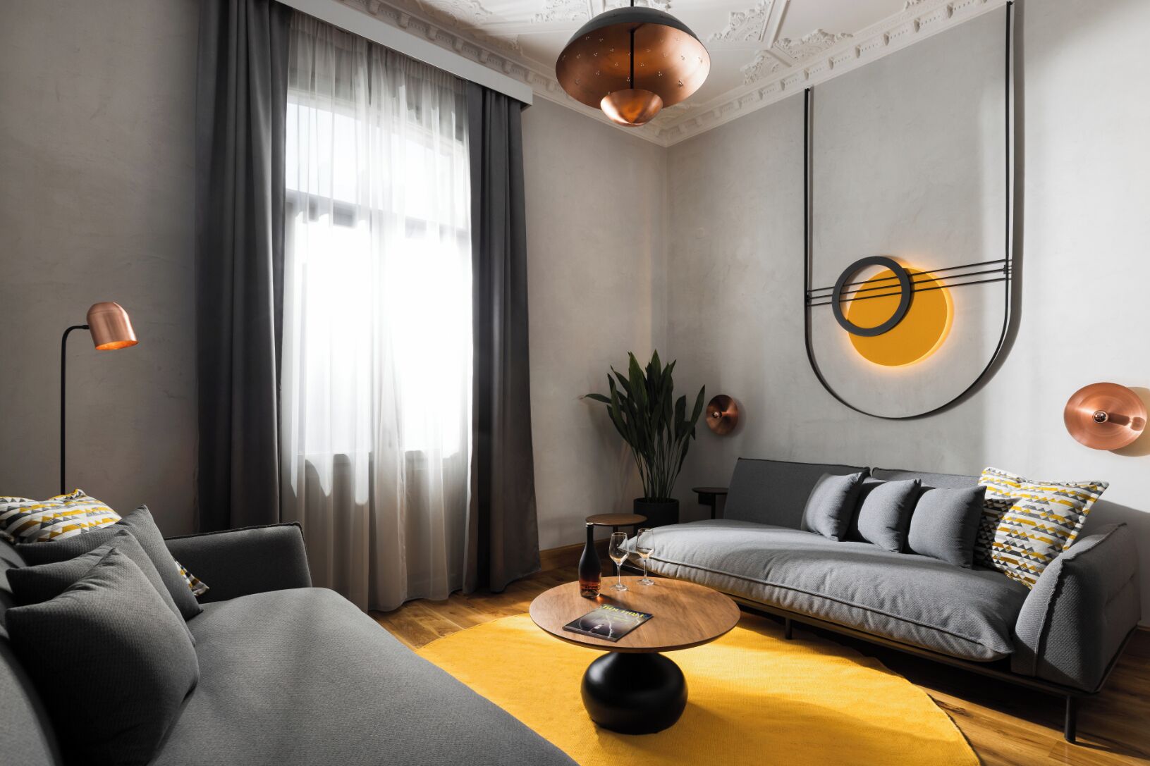 Een moderne woonkamer met een grijze bank, een ronde salontafel en een groot raam met dunne gordijnen.