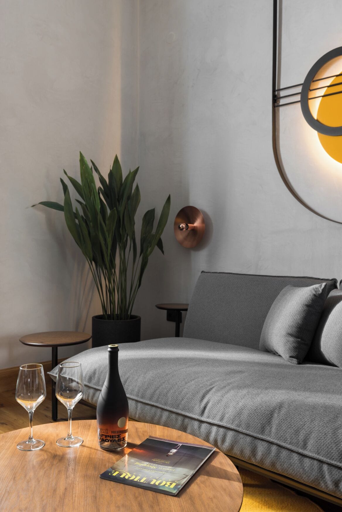 Un salon moderne avec un canapé gris, une table basse ronde sur laquelle se trouvent une bouteille et des verres, une plante en pot et une lampe fixée au mur.
