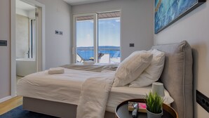 A bedroom with a bed, a bedside table with a lamp, a potted plant, and a view of the sea through large windows.