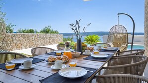 A breakfast setup with a view of the sea, including a table with plates, cups, and a hanging chair.