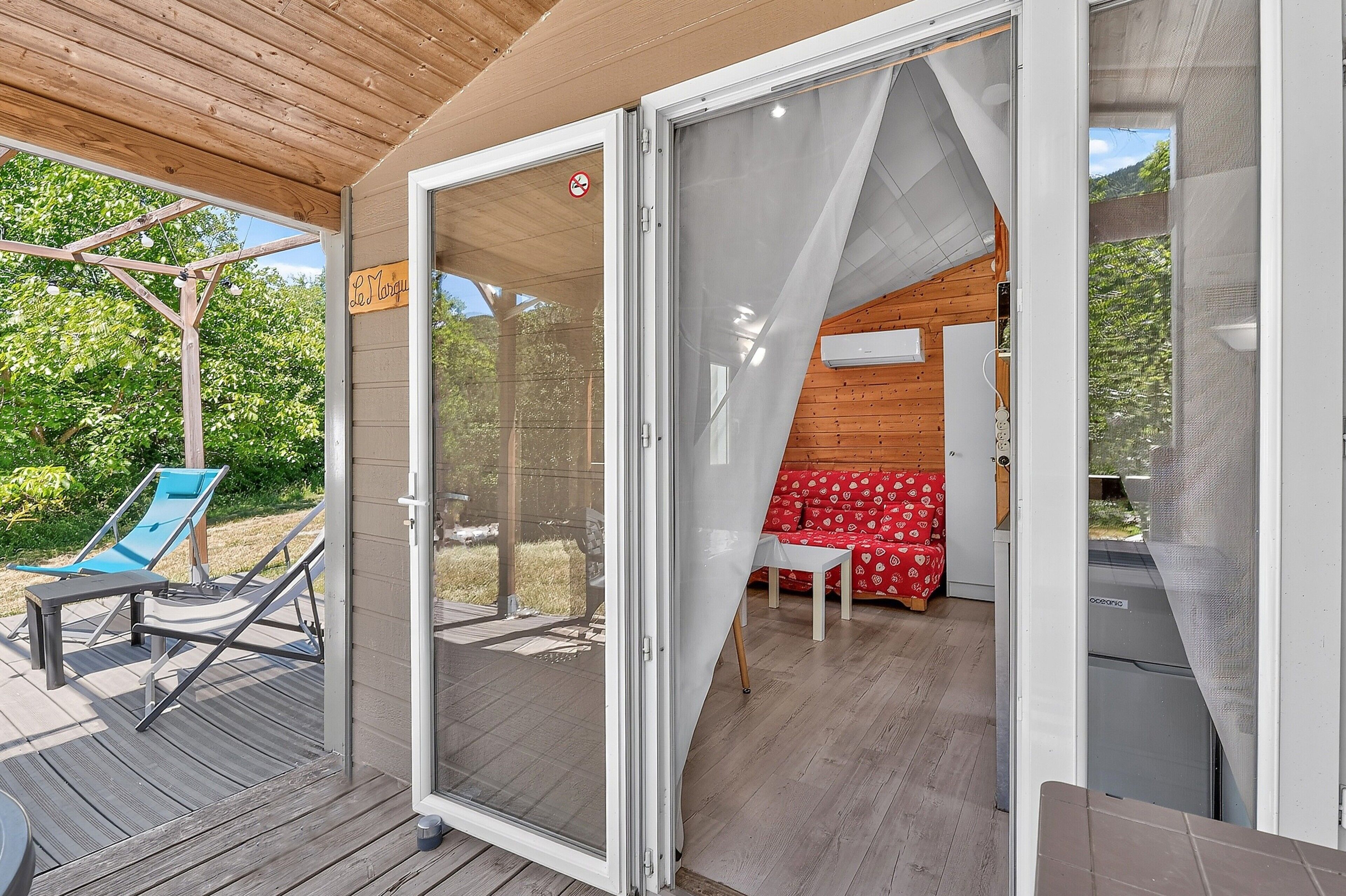 Un espace extérieur moderne avec une terrasse en bois, une porte vitrée donnant sur une pièce où se trouve un canapé rouge à motifs et une petite table, ainsi qu’une enseigne indiquant « Le Marigot ».