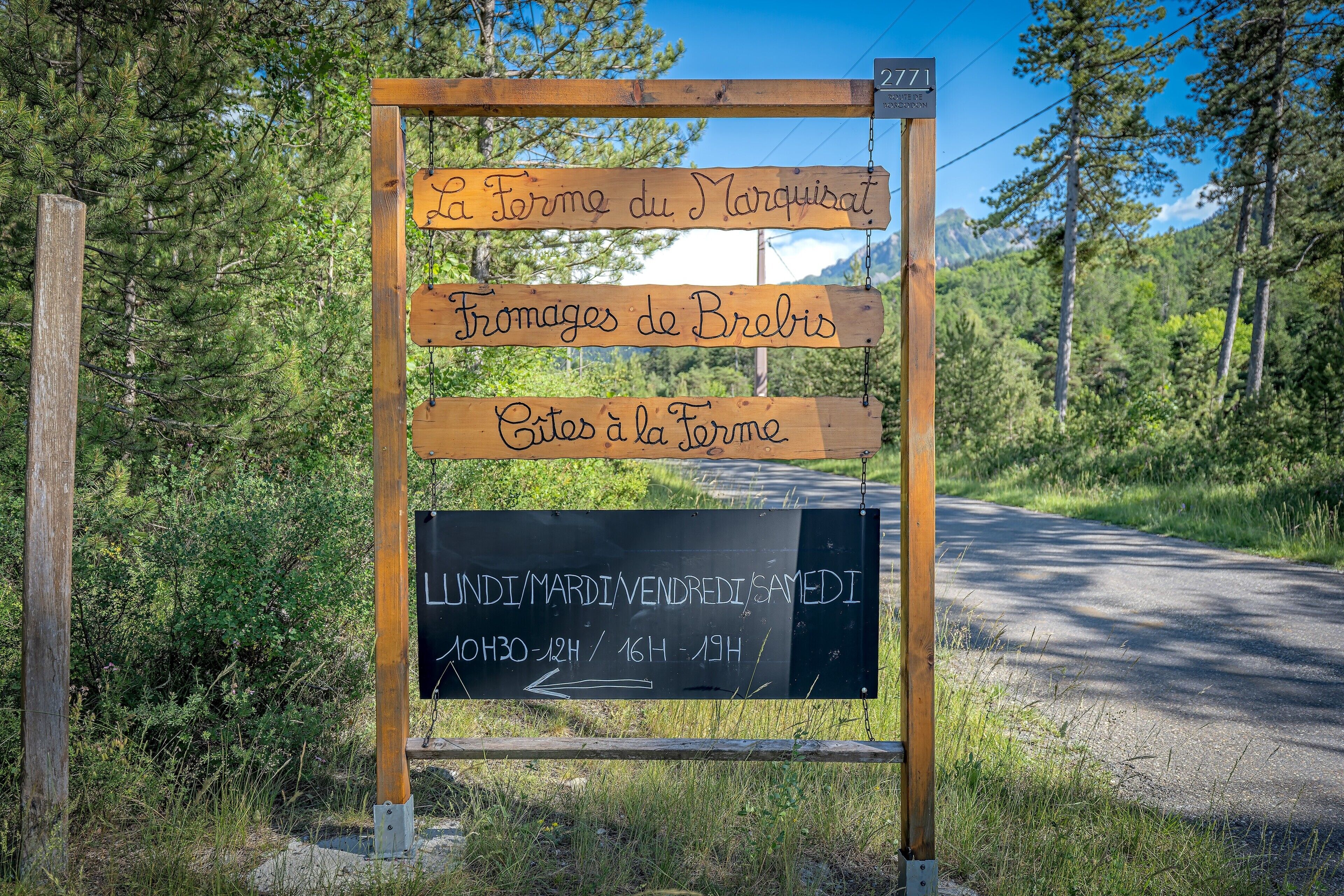 A wooden signpost with text in French, indicating directions to 'La Ferme du Marquisat,' 'Fromages de Brebis,' and 'Cites à la Ferme.'