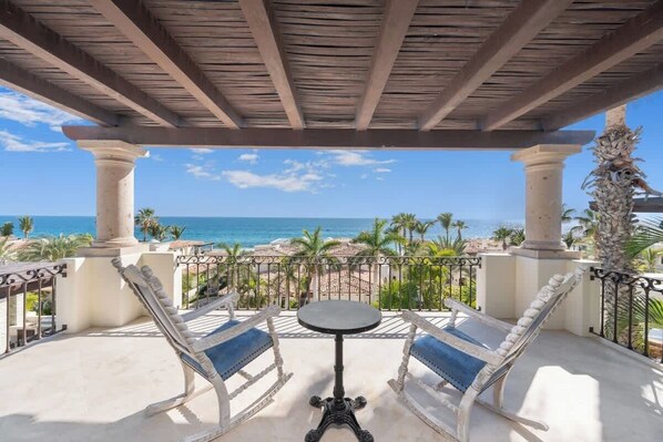 A covered outdoor seating area with two white cushioned chairs and a small table, overlooking a beach with palm trees and the ocean.