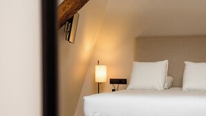 Ein Schlafzimmer mit schrägen Wänden, einem Bett mit weißer Bettwäsche, einer Nachttischlampe und einer an der Wand befestigten Uhr.