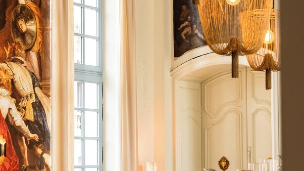 Ein Restaurantinneres mit runden Tischen, gepolsterten Sitzgelegenheiten und einem großen Gemälde im klassischen Stil an der Wand.